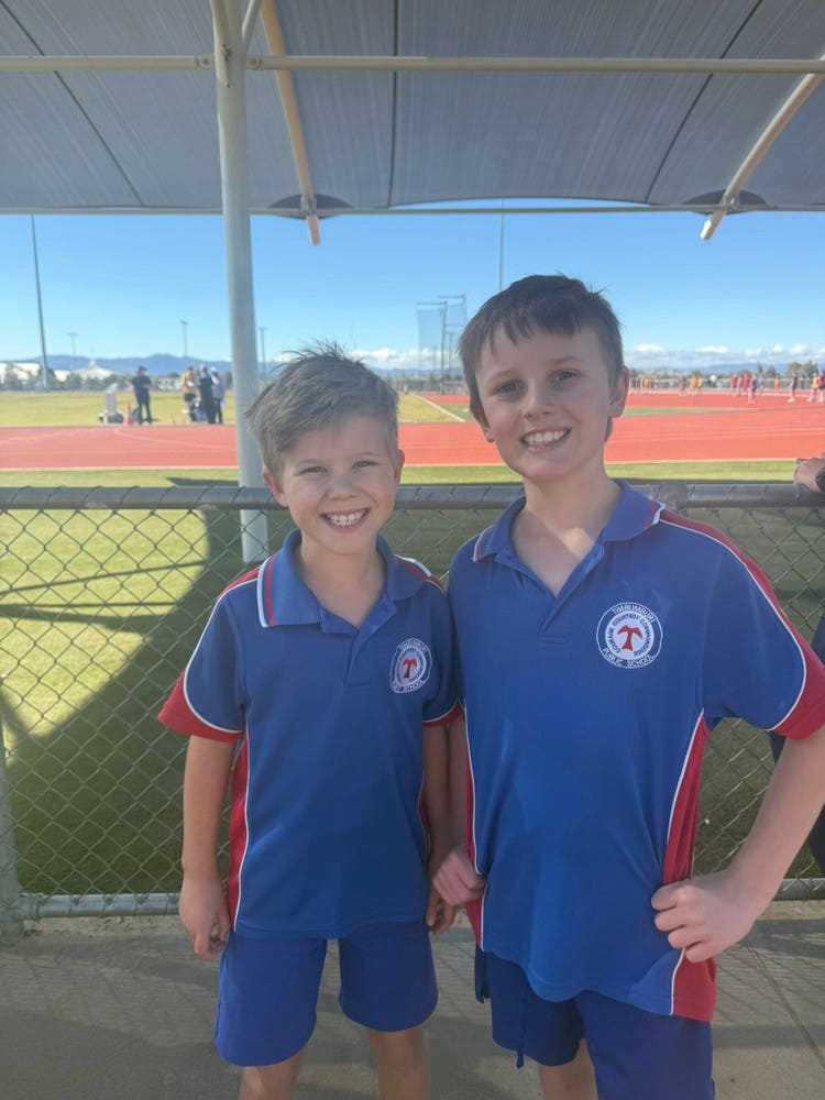 Two boys in blue and red school sports uniforms are standing together at an outdoor athletics track, smiling and ready to participate in sporting events. The sunny sky and field can be seen in the background.