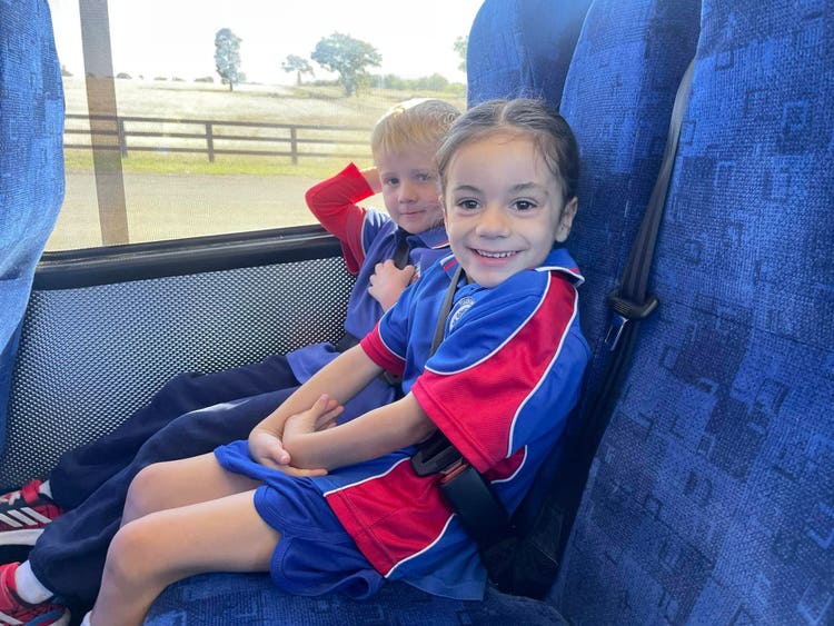 Two students in blue and red school uniforms are seated on a bus, wearing seatbelts, looking out the window at rural scenery during a school excursion.