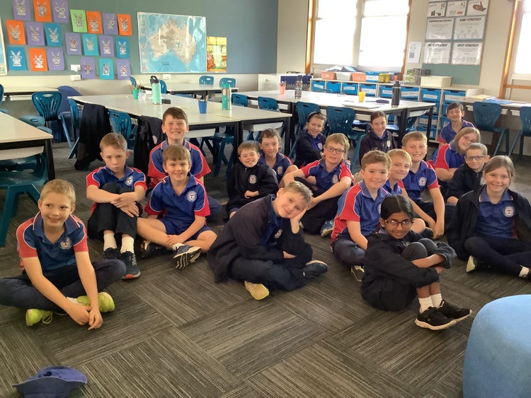 A group of students in blue and red school uniforms are sitting together on the classroom floor, smiling and relaxed, with desks, chairs, and colorful artwork visible in the background.