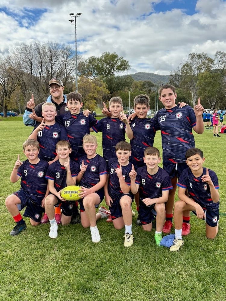 A boys' sports team is posing together on a grassy field, wearing matching navy uniforms with numbers and a school crest. They are holding a football, many with raised index fingers to celebrate a victory or achievement, with trees and a blue sky in the background.