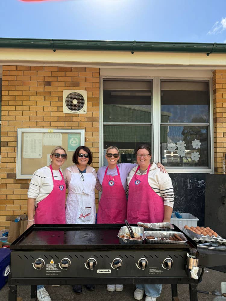 Four adults wearing bright pink aprons are standing outside next to a large barbecue grill, preparing food for a school event, with a brick wall and windows in the background.