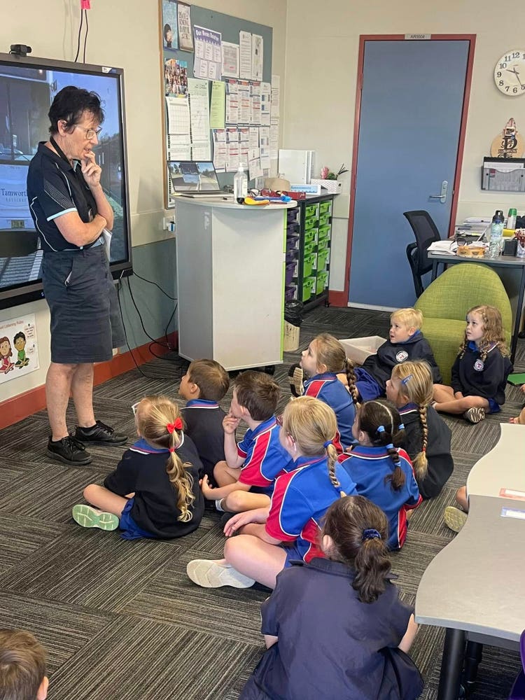 A classroom scene shows a group of students in blue and red uniforms sitting on the floor, attentively listening to a teacher who is standing at the front of the room near a screen and desk.